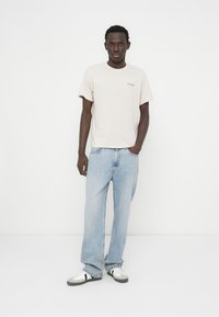 Ανοιχτό ροζ βαμβακερό μπλουζάκι με μικρό κεντημένο λογότυπο, συνδυασμένο με ανοιχτό μπλε, φαρδιά τζιν και μαύρα και λευκά αθλητικά παπούτσια.