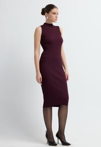Prążkowana, bezrękawna sukienka midi w ciemnym burgundzie z wysokim dekoltem. Noszona z czarnymi szpilkami i przezroczystymi czarnymi rajstopami.