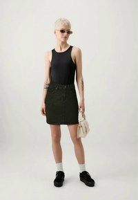 Persona con cabello rubio corto, usando gafas de sol, top sin mangas negro, falda negra con tachuelas, calcetines blancos, zapatos negros, sosteniendo un bolso blanco.