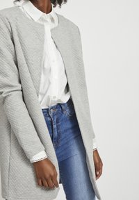 Kvinna klädd i en vit skjorta med knappar, blå jeans och en strukturert ljusgrå öppen cardigan, stående mot en enkel bakgrund.