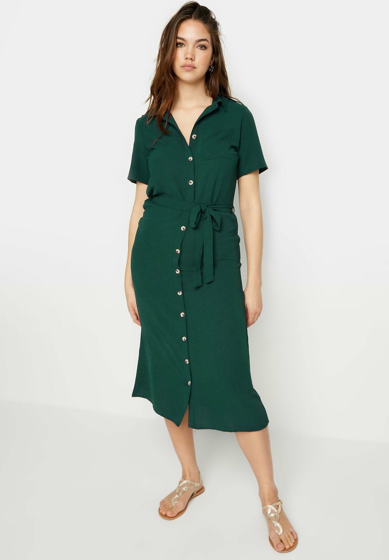 Une robe boutonnée vert foncé en tissu doux, avec une ceinture à la taille et des manches courtes, comportant une fente et des boutons visibles sur le devant.