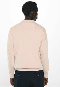 Hellbeige Pullover mit langen Ärmeln, gerippten Bündchen und Saum. Kragen-Design mit weicher Textur. Über dunklen Marineblauen Hosen getragen.