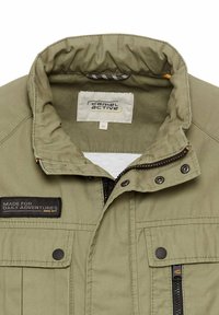 Veste vert olive avec un col haut, présentant une fermeture éclair, un tissu texturé, un patch logo et deux poches poitrine avec des boutons-pression et des fermetures éclair.
