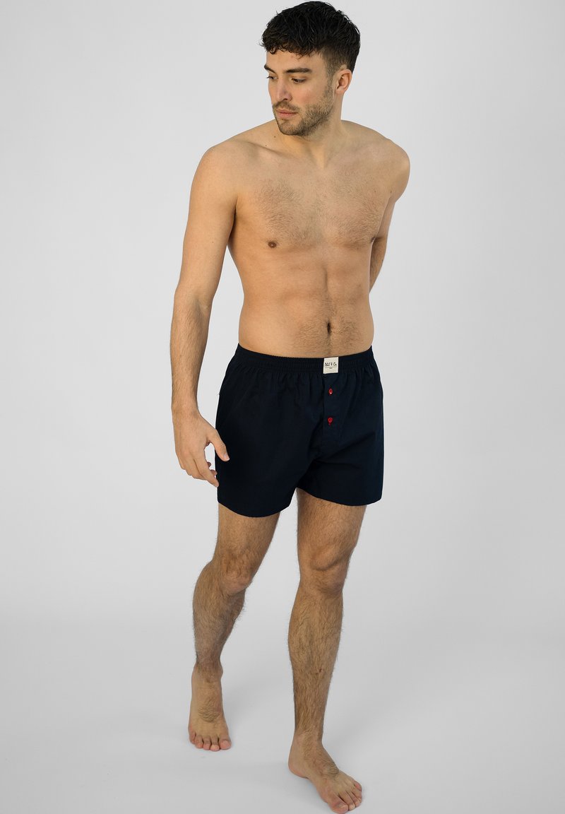 Donkerblauwe boxershorts gemaakt van zachte stof, voorzien van een elastische tailleband en twee rode knopen aan de voorkant voor sluiting.