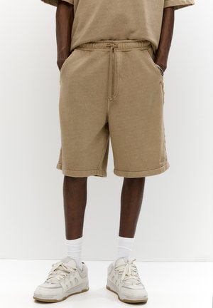 Shorts en mélange de coton beige avec une taille élastique, des poches latérales et des ourlets retroussés, associés à des baskets gris clair et des chaussettes blanches.