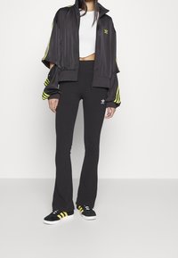 Svart kort jacka med neongula ränder, i kombination med svarta utsvängda byxor som har en Adidas-logotyp, samt svarta skor med gula accenter.