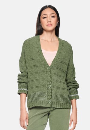 Frau trägt eine lockere, olivgrüne Strickjacke mit Knöpfen, darunter ein hellrosa Oberteil, passende olivgrüne Hose, steht vor weißem Hintergrund.