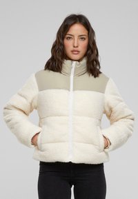 Veste matelassée crème et beige en polaire douce, dotée d'un col haut, d'une fermeture éclair à l'avant et de poches latérales dans un design matelassé.