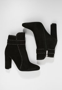 Botines de ante negro con un alto tacón bloque, que presentan detalles de tachuelas y un cierre de cremallera lateral. Textura suave en toda su superficie.