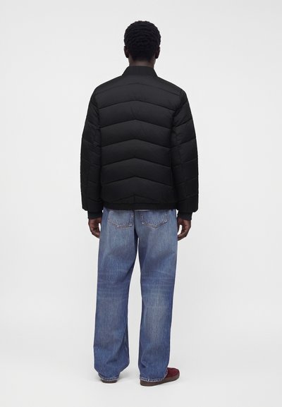 Homme debout, de dos, portant une veste matelassée noire, un jean bleu ample et des chaussures rouges sur un fond blanc uni.