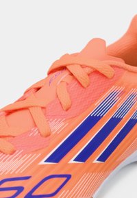 Lyse orange fodboldsko med en struktureret overdel, der har lilla og hvide striber, samt fremtrædende snørebånd for en sikker pasform.