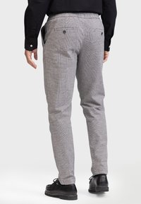 Man in grijze geruite broek, zwart shirt en zwarte leren schoenen, van achteren gezien tegen een effen achtergrond.