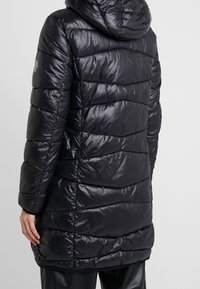 Een persoon in een glanzende zwarte gewatteerde pufferjack met een capuchon, gezien van achteren, met horizontale stiksels en een mouwlogo.