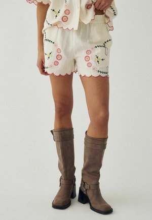 Osoba koja nosi bijele šorceve s ružičastim vezom cvijeća i nazubljenim rubovima, uparene s smeđim čizmama sa srednjim potpeticama do pola lista.