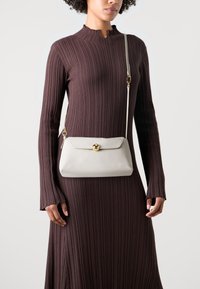 Bílá kožená kabelka s předním uzávěrem a zlatým kováním, nošená přes rameno. Modelka v hnědých žebrovaných šatech s dlouhým rukávem stojí před jednoduchým pozadím.