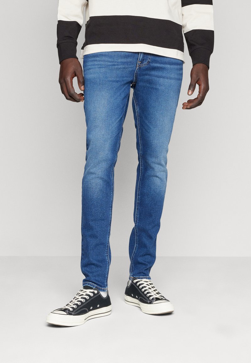 Vaqueros de mezclilla azul ajustados con un acabado desgastado, cinco bolsillos y zapatillas negras a juego con detalles en blanco.