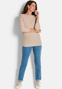 Beige gestrickter Pullover mit Dreiviertelärmeln, kombiniert mit hellblauen, umgeschlagenen Jeans und beigen Turnschuhen, vor einem weißen Hintergrund.