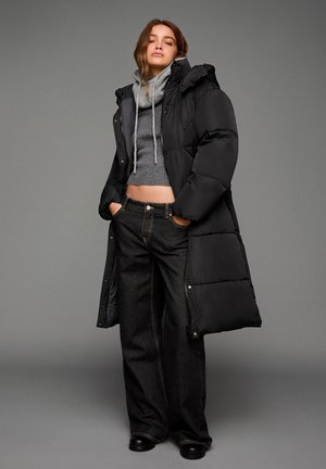 Črn puhasti plašč s kapuco, sivi pleteni pulover in široke črne kavbojke. Gladek material, dizajn do kolen in srebrni dodatki.