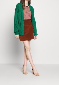 Grüne Strickjacke mit strukturiertem Muster, getragen über einem bordeauxfarbenen gemusterten Oberteil und einem braunen Cordrock. Beige High-Heels.