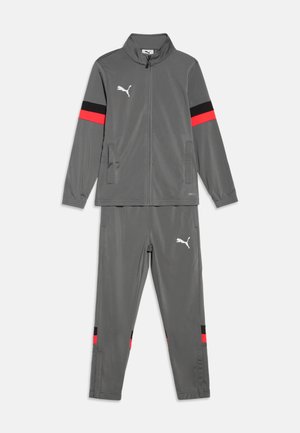 Γκρι φόρμα Puma με ζακέτα με φερμουάρ και παντελόνι, με κόκκινες και μαύρες ρίγες στα μανίκια και στα πόδια, και λευκά λογότυπα Puma στο στήθος και στο μηρό.