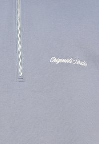 Jasnoszary pulower z suwakiem do ćwiartki wykonany z miękkiej tkaniny, z białym haftowanym napisem „Originals Studio” wzdłuż dolnej części przodu.