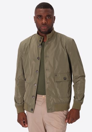 Uomo con giacca verde oliva abbottonata sopra camicia verde scuro e pantaloni beige, in piedi davanti a uno sfondo chiaro e uniforme.