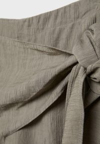 Close-up van beige stof met getextureerde verticale lijnen, in een knoop gestrikt ter hoogte van de taille, met gedetailleerde stiksels en plooien in de stof.