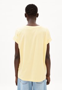 T-shirt di cotone gialla con vestibilità morbida, maniche corte arrotolate e collo rotondo, mostrata da dietro su uno sfondo neutro.