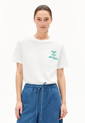 Woman with pulled-back hair wearing a white t-shirt with green text "Made to make a difference" and blue denim drawstring pants.
