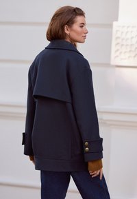 Marineblauwe oversized jas met een achterkantklep, grote revers, gouden knoopaccenten op de manchetten en een geribbelde bruine voering in de mouwen.