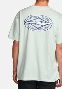 Lyse mintgrøn t-shirt i bomuldsstof, med et blåt grafisk logo på ryggen, der har teksten "BILLABONG BOARDSHORTS" og et bølgedesign.