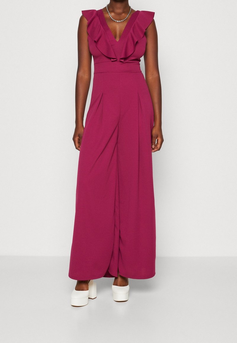 Femme portant une combinaison magenta sans manches avec un col en V à volants et un pantalon à jambes larges, associée à des talons compensés blancs et un collier en chaîne argenté.