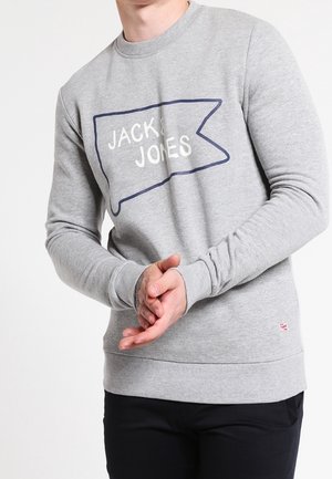 Man draagt een lichtgrijze trui met het logo "JACK & JONES", zwarte broek, staat met gevouwen handen voor zich, effen witte achtergrond.