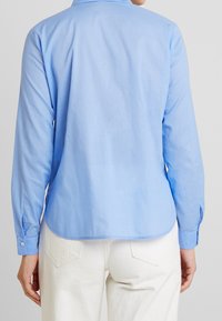 Camicia a maniche lunghe blu chiaro con orlo arrotondato, realizzata in tessuto morbido. Presenta polsini con bottoni e un colletto classico. Indossata con pantaloni bianchi.