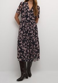Dunkelblaues Kleid mit kurzen Ärmeln, das ein pinkes Blättermuster zeigt, einen V-Ausschnitt hat und einen gestuften Rock besitzt. Kombiniert mit glänzenden, braunen Stiefeln bis zum Knie.