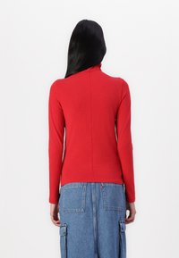Rød langermet genser med høy hals og en tettsittende design. Glatt bomullsstoff, minimal sømdetalj, og høy hals.