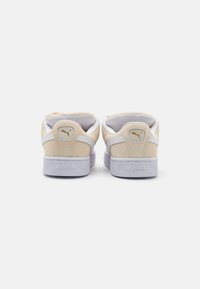 Baskets en suède beige avec des accents en cuir blanc, semelle blanche plate et logo Puma doré sur le talon. Design épuré, style minimaliste.