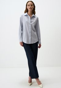 Camicia button-up di un blu chiaro con sottili righe verticali, maniche lunghe e colletto classico, abbinata a pantaloni in denim scuro e tacchi beige.