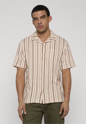 Mężczyzna ubrany w kremową koszulę z krótkim rękawem z pionowymi brązowymi i czerwonymi paskami, zapiętą z przodu na guziki, z kieszenią na piersi oraz oliwkowozielone spodnie.