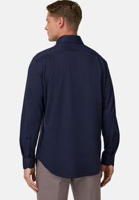 Muž v tmavomodrej košeli s dlhým rukávom a svetlých hnedých nohaviciach, zobrazený zozadu na bielom pozadí.