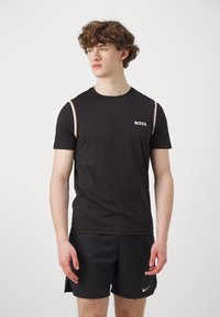 Camiseta atlética negra de manga corta, con un logo blanco de "BOSS" y rayas de hombro en un tono beige que contrastan. Tela suave y transpirable.