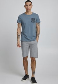 Blå bomulds-t-shirt med en sort mønstret lomme, parret med grå shorts. Model i sorte sneakers, står foran en neutral baggrund.