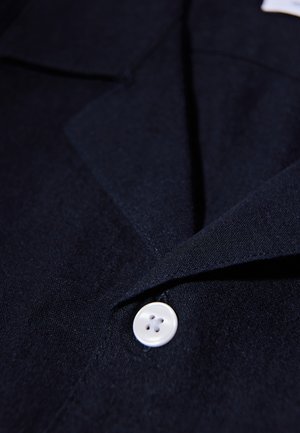 Nahaufnahme einer dunkelroten Stoffjacke mit einem weißen Vierlochknopf, der nahe am umgeschlagenen Kragen befestigt ist.