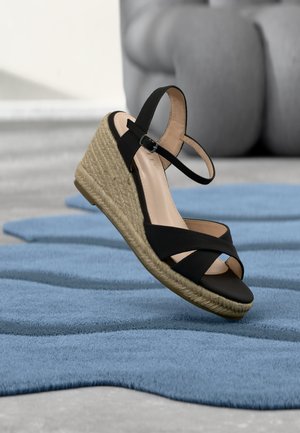 Sandale noire à talon compensé avec plateforme en jute tressé et bride à la cheville reposant sur une surface bleue texturée à l'intérieur.