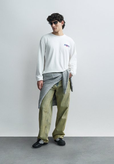 Vīrietis stāv ikdienišķā apģērbā, valkājot melnas saulesbrilles, baltu garroku kreklu, dzeltenzaļas bikses, ap vidukli sasietu pelēku džemperi un melnas sporta kurpes.