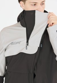 Veste légère en noir et gris. Présente un col haut, des poches zippées et des détails de ventilation en maille. Texture douce et lisse.