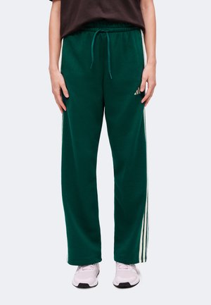 Personne portant un pantalon de jogging vert foncé avec des bandes blanches sur les côtés et des baskets blanches, debout devant un fond uni.
