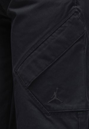 Black cargo pants with a smooth texture, featuring a side pocket and a stitched Air Jordan logo in black on the thigh.