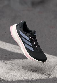 Zwarte sportieve schoen met een gestructureerde bovenkant, drie grijze strepen, roze accenten en een lichtgewicht zool, ontworpen voor prestaties en comfort.