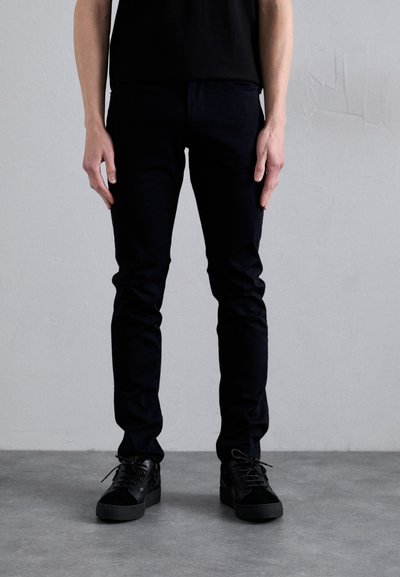 Pantaloni neri aderenti realizzati in tessuto elasticizzato, caratterizzati da una texture liscia e un design elegante, abbinati a scarpe nere con lacci.